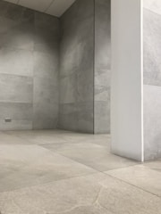 A room with minimalist design, featuring light gray stone tiles covering the floor and walls. The space is empty, with a single white pillar extending vertically from the floor to the ceiling. The lighting is soft and even, adding to the stark and modern architectural feel.