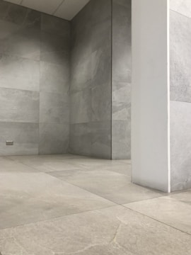 A room with minimalist design, featuring light gray stone tiles covering the floor and walls. The space is empty, with a single white pillar extending vertically from the floor to the ceiling. The lighting is soft and even, adding to the stark and modern architectural feel.