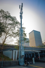A tall telecommunications tower stands against a city backdrop. Nearby, a modern building with a grid of windows is visible. A highway overpass runs through the middle of the scene, with two people sitting on the ground near the base of the tower. Trees with green foliage partially frame the view on the left.