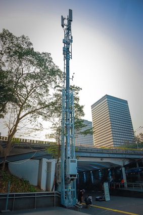 A tall telecommunications tower stands against a city backdrop. Nearby, a modern building with a grid of windows is visible. A highway overpass runs through the middle of the scene, with two people sitting on the ground near the base of the tower. Trees with green foliage partially frame the view on the left.