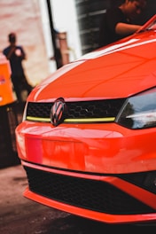 A vibrant orange Volkswagen car is prominently displayed. The focus is on the front, highlighting the logo and the sleek design. In the background, two people are faintly visible, one of whom appears to be engaging in some form of work near the vehicle.