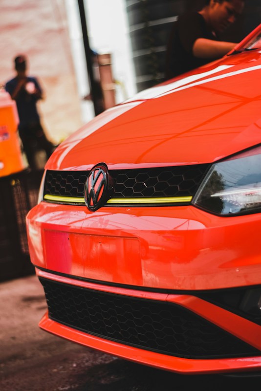 A vibrant orange Volkswagen car is prominently displayed. The focus is on the front, highlighting the logo and the sleek design. In the background, two people are faintly visible, one of whom appears to be engaging in some form of work near the vehicle.
