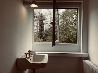A minimalistic interior scene featuring a wall-mounted sink with vintage-style taps, placed beneath a light fixture. An open window reveals a view of trees and foliage outside, allowing natural light to fill the space. A radiator is visible on the right side of the image.
