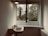 A minimalistic interior scene featuring a wall-mounted sink with vintage-style taps, placed beneath a light fixture. An open window reveals a view of trees and foliage outside, allowing natural light to fill the space. A radiator is visible on the right side of the image.