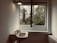 A minimalistic interior scene featuring a wall-mounted sink with vintage-style taps, placed beneath a light fixture. An open window reveals a view of trees and foliage outside, allowing natural light to fill the space. A radiator is visible on the right side of the image.