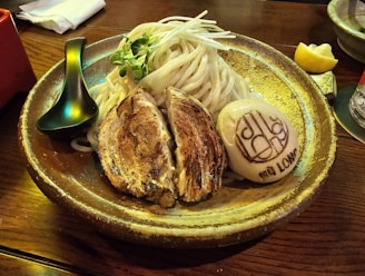 A ceramic bowl contains a carefully arranged dish featuring sliced grilled pork, creamy-colored noodles garnished with green sprouts, and a boiled egg with a decorative imprint. A black soup spoon rests on the bowl's edge. A lemon wedge and a red-labeled drink appear in the background on a wooden table.