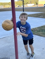 A young boy, smiling broadly, stands outdoors holding a tetherball attached to a red pole. He is wearing a blue shirt, black shorts, and gray sneakers. The background features a paved area, grass, several parked vehicles, including yellow school buses, and trees.