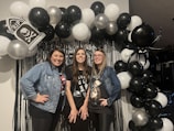 Three women are posing in front of a festive balloon arch, which includes black, white, and silver balloons. The arch features elements like a tombstone with 'RIP', skull designs, spider patterns, and a large black spider balloon. The women are dressed casually and appear happy, standing before a backdrop with metallic fringe and additional decorations.