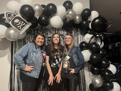 Three women are posing in front of a festive balloon arch, which includes black, white, and silver balloons. The arch features elements like a tombstone with 'RIP', skull designs, spider patterns, and a large black spider balloon. The women are dressed casually and appear happy, standing before a backdrop with metallic fringe and additional decorations.