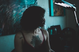 A person with curly hair is sitting under a lamp, partially illuminated, with a visible tattoo on their shoulder and upper chest. The room is dimly lit with abstract artwork and equipment in the background.