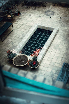 A tiled courtyard features a rectangular water feature with a grid pattern at the bottom. Red and white flowers in pots are placed at one end, adding a touch of color. Benches and potted plants are arranged around the courtyard, creating a calm and serene environment.