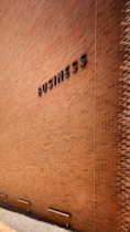 A large brick wall with the word 'BUSINESS' prominently displayed in black letters. The wall is lit by natural daylight, casting gentle shadows along its surface. The ground at the base of the wall has some patches of grass and is bordered by a concrete sidewalk. There are three metal fixtures attached to the lower part of the wall.