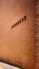 A large brick wall with the word 'BUSINESS' prominently displayed in black letters. The wall is lit by natural daylight, casting gentle shadows along its surface. The ground at the base of the wall has some patches of grass and is bordered by a concrete sidewalk. There are three metal fixtures attached to the lower part of the wall.