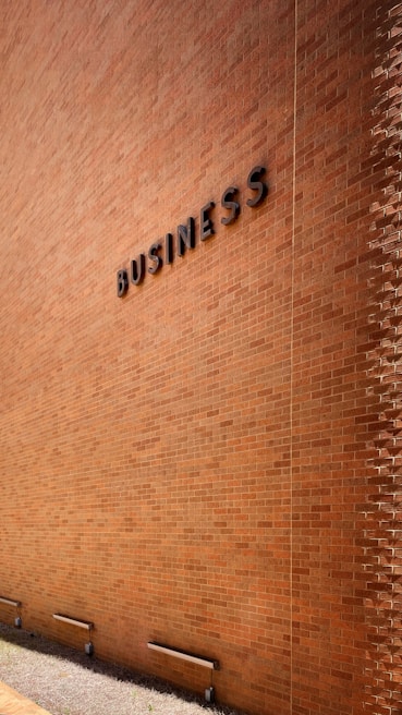 A large brick wall with the word 'BUSINESS' prominently displayed in black letters. The wall is lit by natural daylight, casting gentle shadows along its surface. The ground at the base of the wall has some patches of grass and is bordered by a concrete sidewalk. There are three metal fixtures attached to the lower part of the wall.