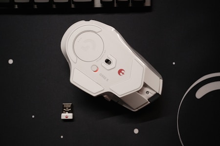A white gaming mouse with an ergonomic design is placed on a black surface. Beside the mouse, there is a small USB receiver. The mouse has some branding, including the model number G502 X.