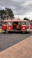 A vintage red Volkswagen van with open side doors, revealing an interior decorated with red seating and a fluffy pillow. The van is parked on a gravel pathway next to a brick-paved area. Overhead, power lines stretch across a cloudy sky, while trees and urban features like streetlights and signs can be seen in the background.