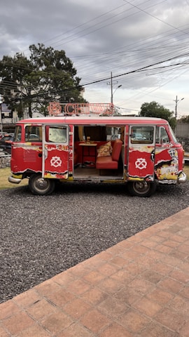 A vintage red Volkswagen van with open side doors, revealing an interior decorated with red seating and a fluffy pillow. The van is parked on a gravel pathway next to a brick-paved area. Overhead, power lines stretch across a cloudy sky, while trees and urban features like streetlights and signs can be seen in the background.