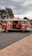 A vintage red Volkswagen van with open side doors, revealing an interior decorated with red seating and a fluffy pillow. The van is parked on a gravel pathway next to a brick-paved area. Overhead, power lines stretch across a cloudy sky, while trees and urban features like streetlights and signs can be seen in the background.