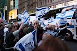 A crowd of people is gathered in the street, holding and waving Israeli flags. The setting appears to be an urban environment, with buildings and buses in the background. The crowd is dense, and the atmosphere seems lively and energetic.