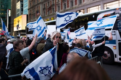 A crowd of people is gathered in the street, holding and waving Israeli flags. The setting appears to be an urban environment, with buildings and buses in the background. The crowd is dense, and the atmosphere seems lively and energetic.