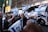 A crowd of people is gathered in the street, holding and waving Israeli flags. The setting appears to be an urban environment, with buildings and buses in the background. The crowd is dense, and the atmosphere seems lively and energetic.
