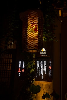 A warmly lit traditional lantern hangs against a dark background. The lantern's mesh pattern allows light to filter through, casting a soft, inviting glow. Below it, there is a dark board with illuminated vertical text lines, accompanied by green leaves draping around.