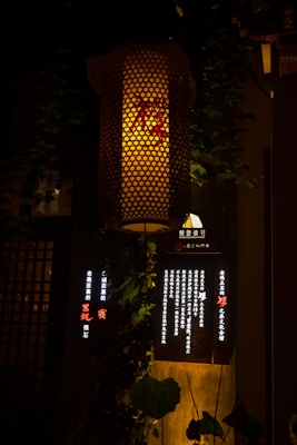 A warmly lit traditional lantern hangs against a dark background. The lantern's mesh pattern allows light to filter through, casting a soft, inviting glow. Below it, there is a dark board with illuminated vertical text lines, accompanied by green leaves draping around.