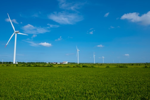 Several wind turbines are scattered across a vast, green countryside under a bright blue sky with a few scattered clouds. A small building is visible in the distance, surrounded by lush greenery.