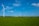 Several wind turbines are scattered across a vast, green countryside under a bright blue sky with a few scattered clouds. A small building is visible in the distance, surrounded by lush greenery.
