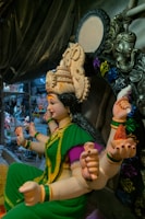 A detailed statue of a deity with multiple arms, adorned in a green and yellow sari with vibrant purple accents. The headdress is elaborately decorated, and the figure is seated in a position suggesting poise and grace. Surrounding the statue are various decorative elements and other sculptures, indicative of a workshop or studio setting.