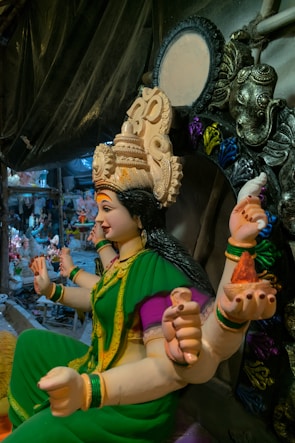 A detailed statue of a deity with multiple arms, adorned in a green and yellow sari with vibrant purple accents. The headdress is elaborately decorated, and the figure is seated in a position suggesting poise and grace. Surrounding the statue are various decorative elements and other sculptures, indicative of a workshop or studio setting.