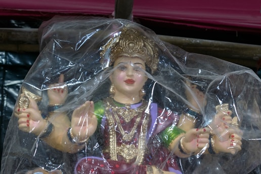 A colorful statue of a goddess with multiple arms is wrapped in transparent plastic. The idol is adorned with intricate jewelry, including a detailed crown, and is richly painted in vibrant colors such as red, purple, and gold.