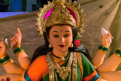 A detailed sculpture of a multi-armed deity adorned with an intricately designed golden headdress and jewelry. The figure is wearing a colorful garment with green, orange, and gold hues. The hands are positioned in various symbolic gestures, holding objects such as a shell and a discus.