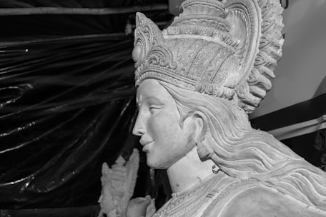 A monochrome image of a detailed sculpture depicting a figure wearing an ornate headdress. The craftsmanship shows intricate patterns on the headpiece, and the figure's facial features are finely carved. The background is dark, contrasting with the light-toned sculpture.