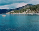 A serene lake surrounded by dense pine forests and rocky hills. The water is a clear shade of blue, reflecting the overcast sky. Small rock formations are scattered near the shore, and the distant mountains are partially shrouded in mist, adding to the tranquil and somewhat mysterious atmosphere.