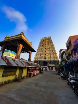 A towering ancient temple structure stands majestically against a clear blue sky. The architecture features intricate carvings and is flanked by smaller structures. Shops and motorcycles line the pathway leading to the temple, adding a sense of liveliness to the scene.