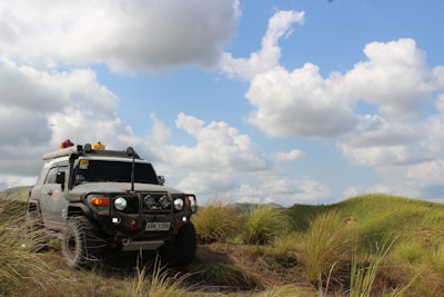 A rugged off-road vehicle is parked on a grassy field with rolling hills in the background. The sky is partly cloudy, with large, white clouds scattered across a bright blue sky. The vehicle is equipped for off-roading, featuring equipment such as a roof rack, auxiliary lights, and a snorkel.