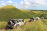 A group of off-road vehicles is traversing a scenic, grassy landscape with rolling hills and scattered clouds in the sky. The terrain is lush and green, suggesting a natural, unspoiled environment. A person in a blue outfit is seen near one of the vehicles, adding a sense of adventure and exploration to the scene.