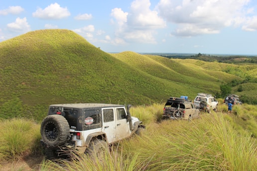 A group of off-road vehicles is traversing a scenic, grassy landscape with rolling hills and scattered clouds in the sky. The terrain is lush and green, suggesting a natural, unspoiled environment. A person in a blue outfit is seen near one of the vehicles, adding a sense of adventure and exploration to the scene.