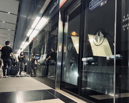 A modern metro station platform featuring glass doors that separate the platform from the train tracks. Several people are standing and walking, some with luggage, suggesting a busy environment. The lighting is bright, and there are reflections visible on the glass surfaces.
