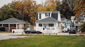 A suburban neighborhood scene featuring several houses with well-maintained lawns and a variety of trees displaying fall foliage. In the foreground, a sporty black car is parked in a driveway alongside two other vehicles. The houses have different architectural styles, including a two-story house with a white picket fence and a one-story house with a front porch.