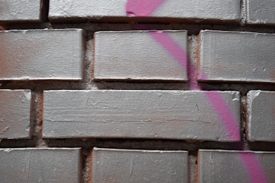 Rectangular bricks with a rough texture are arranged in a neat pattern. A streak of pink spray paint crosses over several bricks, adding a vibrant contrast to the muted color of the bricks.
