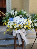 A large and vibrant floral arrangement features a mix of white, yellow, and blue flowers, including roses, daisies, and hydrangeas. The arrangement is complemented by lush green foliage and secured with a wide white ribbon tied in a bow. It is placed on a wooden stand in an outdoor setting against a textured beige wall, with part of a building entrance visible in the background.