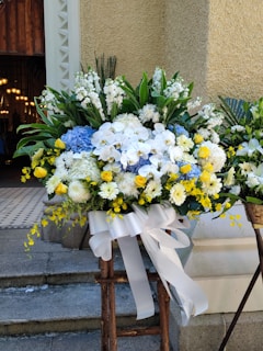 A large and vibrant floral arrangement features a mix of white, yellow, and blue flowers, including roses, daisies, and hydrangeas. The arrangement is complemented by lush green foliage and secured with a wide white ribbon tied in a bow. It is placed on a wooden stand in an outdoor setting against a textured beige wall, with part of a building entrance visible in the background.