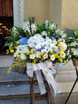 A large and vibrant floral arrangement features a mix of white, yellow, and blue flowers, including roses, daisies, and hydrangeas. The arrangement is complemented by lush green foliage and secured with a wide white ribbon tied in a bow. It is placed on a wooden stand in an outdoor setting against a textured beige wall, with part of a building entrance visible in the background.