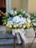 A large and vibrant floral arrangement features a mix of white, yellow, and blue flowers, including roses, daisies, and hydrangeas. The arrangement is complemented by lush green foliage and secured with a wide white ribbon tied in a bow. It is placed on a wooden stand in an outdoor setting against a textured beige wall, with part of a building entrance visible in the background.