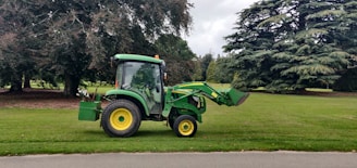 A green tractor with a front loader attachment is parked on a grassy area next to a paved path. Large trees with dense foliage are visible in the background, creating a serene outdoor setting.