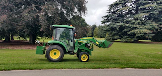 A green tractor with a front loader attachment is parked on a grassy area next to a paved path. Large trees with dense foliage are visible in the background, creating a serene outdoor setting.
