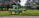 A green tractor with a front loader attachment is parked on a grassy area next to a paved path. Large trees with dense foliage are visible in the background, creating a serene outdoor setting.