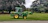 A green tractor with a front loader attachment is parked on a grassy area next to a paved path. Large trees with dense foliage are visible in the background, creating a serene outdoor setting.